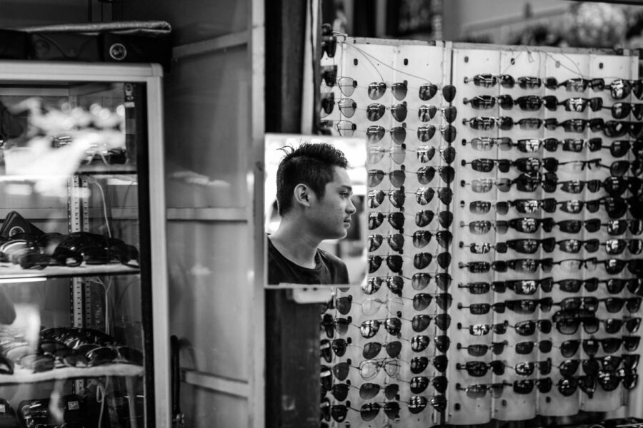 Petaling Street hawker store in Chinatown - Kuala Lumpur - Malaysia. Street photograph, of a man's face reflected in a mirror used to see the sunglasses on your face.