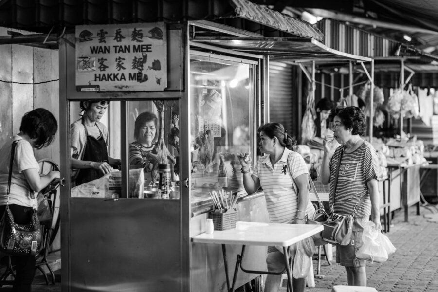 Petaling Street, Chinatown food hawker stall selling wan tan mee, hakka me. Woman customers test tasting the food. Black and white street photo Kuala Lumpur - Malaysia